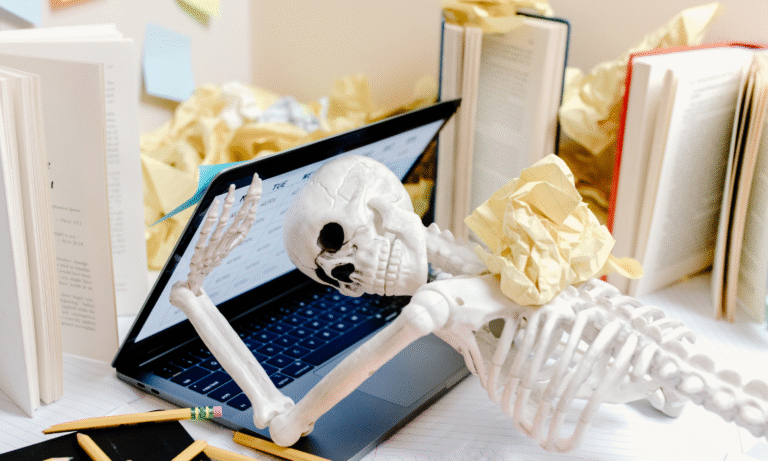 Skeleton slumped over laptop on messy desk with books
